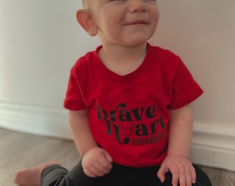 May include: A young child wearing a red t-shirt with the words "brave heart" in black script and a heart graphic. The shirt also includes the hashtag #CHDAWARENESS. The child is also wearing black pants and is sitting on a wooden floor.