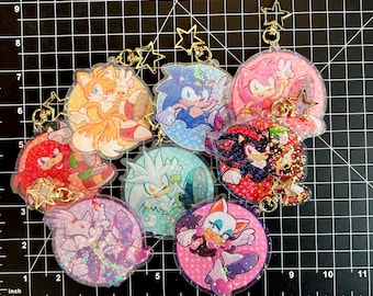 May include: A set of eight round, clear, glitter-filled keychains featuring characters from the Sonic the Hedgehog video game franchise. Each keychain has a gold star charm attached to a gold chain. The characters include Sonic, Tails, Knuckles, Shadow, Amy, Silver, Blaze, and Rouge.