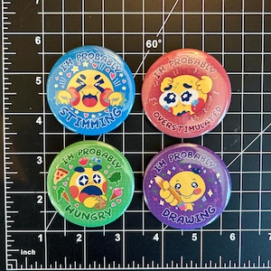 Puede incluir: Cuatro pines de botón de 5,7 cm de diámetro con fondos coloridos y caras de dibujos animados amarillas. El texto en los pines dice "Probablemente estoy estimulando", "Probablemente estoy sobreestimulado", "Probablemente tengo hambre" y "Probablemente estoy dibujando".