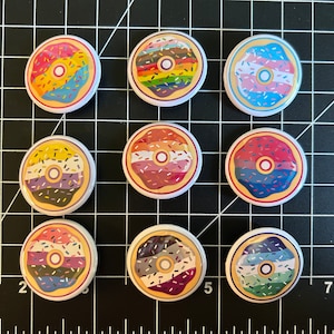 Puede incluir: Nueve botones redondos con pasador con diferentes diseños de donuts con chispas y glaseado en varios colores, incluyendo rosa, azul, amarillo, verde y morado.