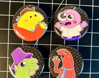 Pode incluir: Quatro botões redondos com fundos de bolinhas pretas e brancas apresentando personagens de desenhos animados. Os personagens são uma criatura amarela vestindo um moletom vermelho, uma criatura rosa vestindo uma camisa branca e calças azuis, uma criatura verde vestindo um chapéu roxo e uma criatura vermelha vestindo uma gravata azul.
