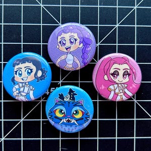 May include: Four round pinback buttons with cartoon illustrations. The buttons feature characters with purple, pink, and blue color schemes. One button has a blue cat design. The buttons are arranged on a black grid.