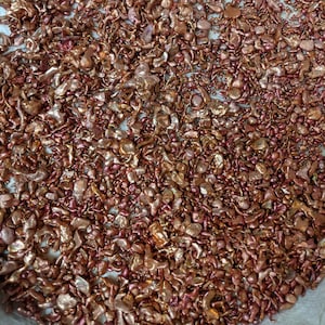 May include: A close-up image of a large quantity of small, irregularly shaped pieces of copper metal. The copper pieces are a reddish-brown color and have a metallic sheen.