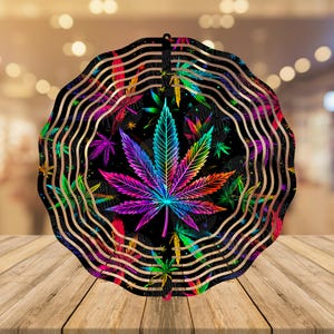 May include: A colorful metal wind spinner with a large, rainbow-colored marijuana leaf in the center. The spinner has a black background with smaller marijuana leaves scattered around the large leaf. The spinner is designed to spin in the wind.