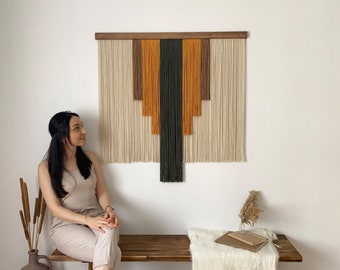 May include: A woman sits on a wooden bench in front of a wall hanging made of yarn. The wall hanging is a geometric design with beige, brown, orange, and black yarn. The bench has a white throw blanket on it.