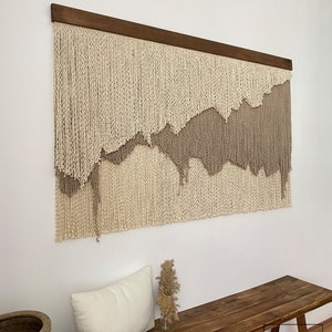 A beige and brown macrame wall hanging with a wooden frame. The hanging features a wavy, abstract design.
