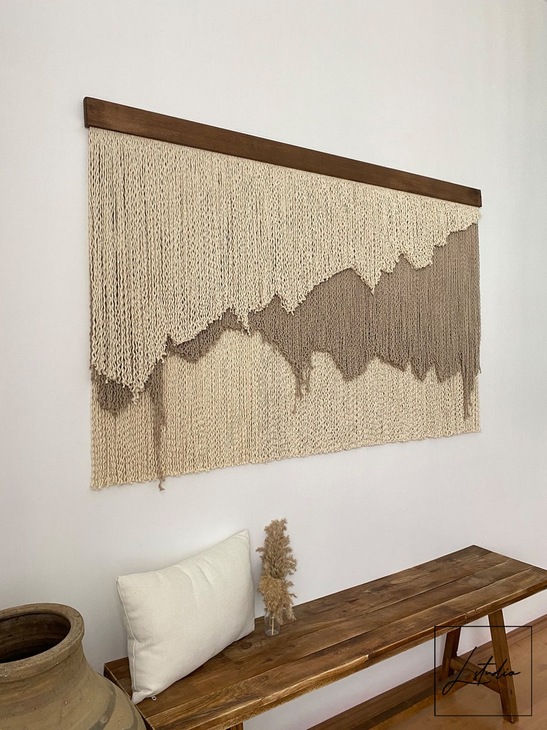 A beige and brown macrame wall hanging with a wooden frame. The hanging features a wavy, abstract design.