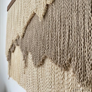 A wall hanging made of woven natural fiber rope with a gradient of beige and brown colors. The rope is woven in a pattern that creates a series of waves or scallops. The wall hanging is mounted on a wooden frame.
