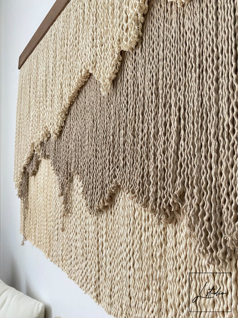 A wall hanging made of woven natural fiber rope with a gradient of beige and brown colors. The rope is woven in a pattern that creates a series of waves or scallops. The wall hanging is mounted on a wooden frame.