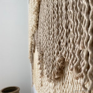 A beige and cream macrame wall hanging with a woven texture. The hanging is made of natural fibers and has a fringed edge.