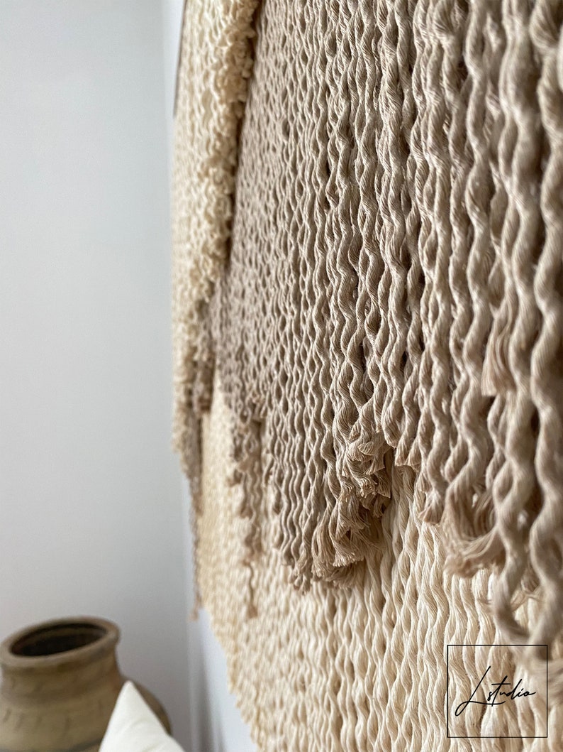 A beige and cream macrame wall hanging with a woven texture. The hanging is made of natural fibers and has a fringed edge.