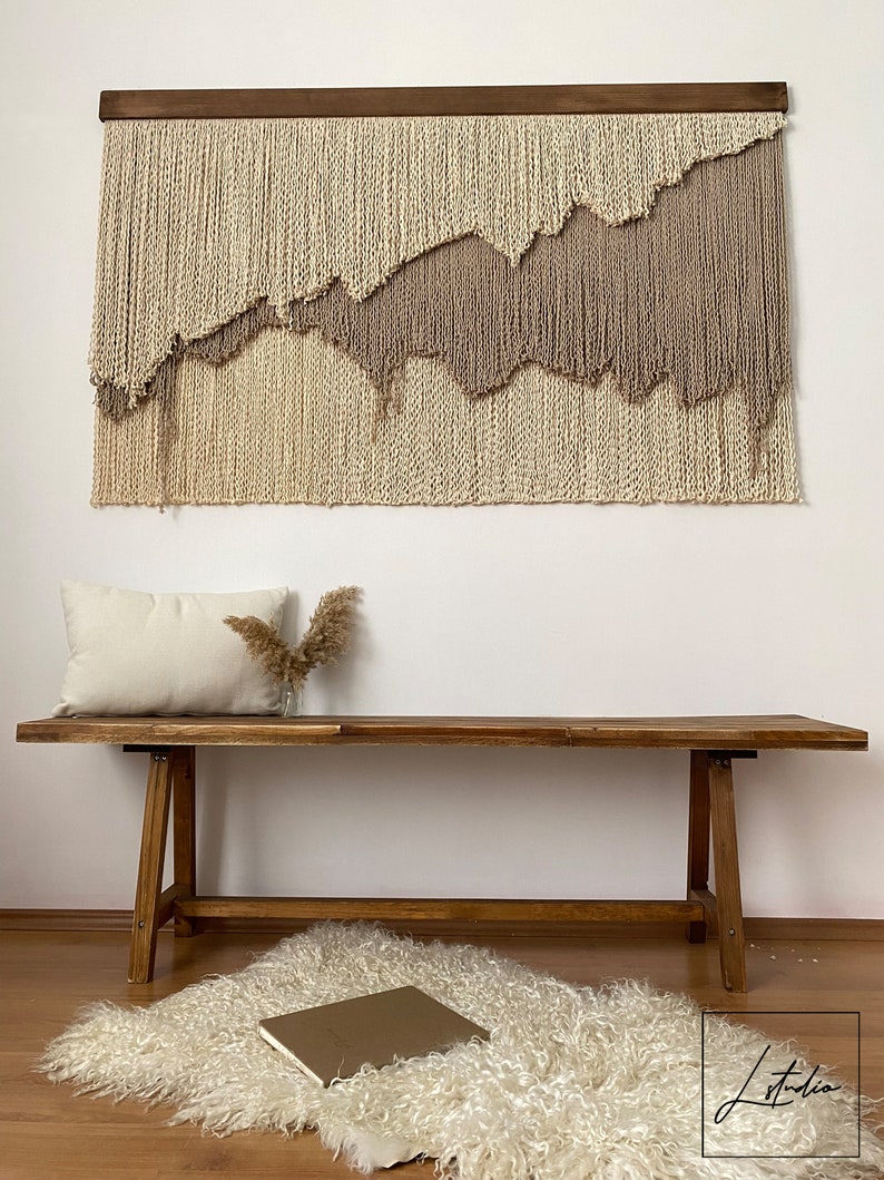 A beige and brown taupe macrame wall hanging with a wooden frame. The hanging is made of yarn and has a wavy, abstract design. The hanging is displayed on a white wall above a wooden bench with a white pillow and a vase of dried pampas grass.