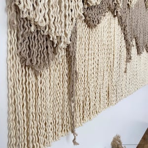 A beige and brown macrame wall hanging with a fringe design. The hanging is made of twisted rope and has a layered, geometric pattern.