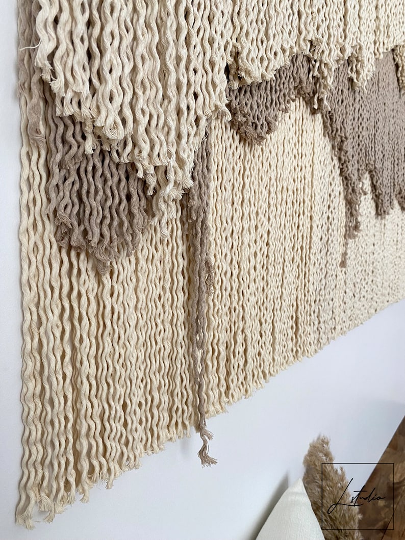 A beige and brown macrame wall hanging with a fringe design. The hanging is made of twisted rope and has a layered, geometric pattern.