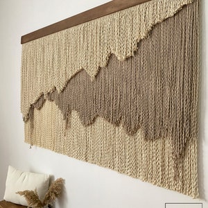 A large wall hanging made of woven yarn with a wood frame. The yarn is in two shades of beige, creating a layered effect. The hanging has a wavy pattern.