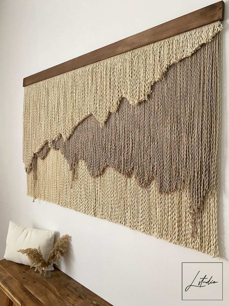 A large wall hanging made of woven yarn with a wood frame. The yarn is in two shades of beige, creating a layered effect. The hanging has a wavy pattern.