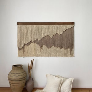 A beige and taupe macrame wall hanging with a wooden dowel at the top. The wall hanging is made of yarn and has a wavy, abstract design. The wall hanging is hanging on a white wall.