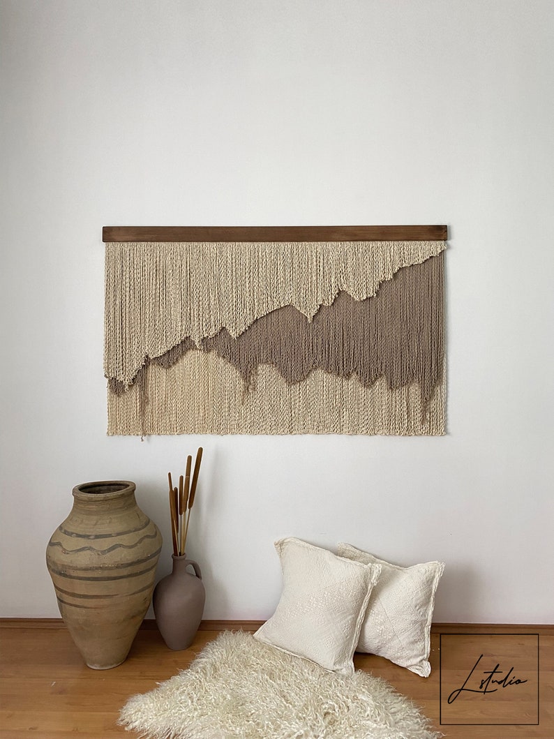 A beige and taupe macrame wall hanging with a wooden dowel at the top. The wall hanging is made of yarn and has a wavy, abstract design. The wall hanging is hanging on a white wall.