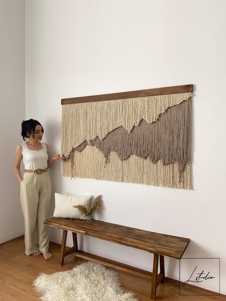 A large beige and brown macrame wall hanging with a wooden frame. The wall hanging is made of yarn and has a wavy, abstract design.