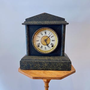 May include: A mantel clock with a dark green faux marble finish and a triangular pediment top. The clock face features a gold-toned circular center with black Roman numerals, two black hands, and two winding holes on a white background.