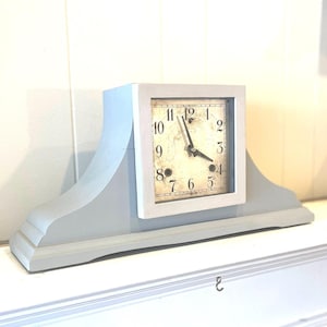 May include: A gray painted mantle clock with a white face and black hands. The clock face has black Roman numerals and a gold-colored background.