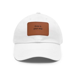 May include: A white baseball cap featuring a rectangular brown patch centred on the front. The patch displays the black text "Print it your way" in a simple font. The cap has a curved brim and a structured crown with stitched eyelets.