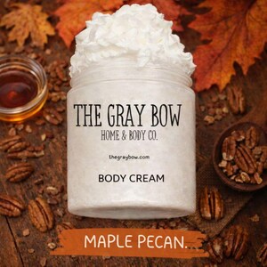 Könnte beinhalten: Ein Glas Körpercreme mit Schlagsahne, beschriftet mit "The Gray Bow Home & Body Co.". Das Etikett lautet auch "Body Cream" und "Maple Pecan". Der Hintergrund umfasst Herbstblätter, Pekannüsse und ein kleines Glas Flüssigkeit.