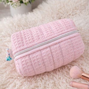 May include: A pale pink rectangular makeup bag with a waffle-weave pattern and a silver zip. The bag rests on a fluffy, cream-coloured surface, with a bouquet of pink roses in the background. Makeup brushes are also visible.