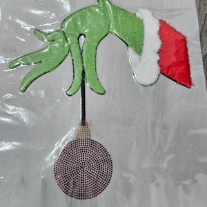 Puede incluir: Una mano verde con un gorro de Papá Noel rojo y blanco sosteniendo un adorno morado cubierto de pedrería. El diseño está sobre un fondo blanco.