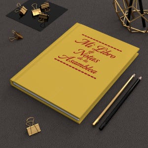 Pode incluir: Um caderno de capa dura na cor amarelo mostarda com o texto Mi Libro de Notas de la Asamblea impresso em vermelho escuro na capa, acompanhado por dois lápis e vários clipes de metal dourado dispostos ao redor do caderno.