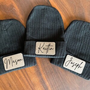 May include: Three black ribbed knit beanies with personalized leatherette patches. The patches are rectangular and feature the names "Mason", "Kaitlin", and "Joseph" in a stylish script font. The beanies are displayed on a wooden surface.