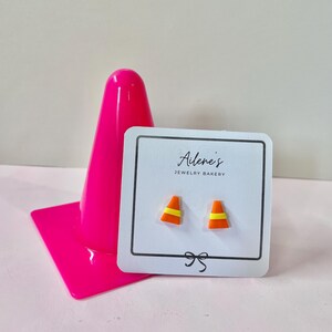 Puede incluir: Un par de pendientes de cono de tráfico naranja y amarillo en una tarjeta blanca con el texto "Ailene's JEWELRY BAKERY". Un cono de tráfico rosa brillante está en el fondo.
