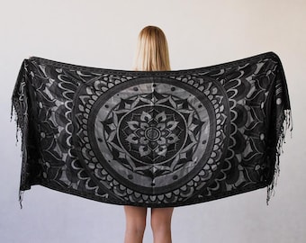 May include: A rectangular black and grey scarf featuring a large, intricate mandala pattern centered on the fabric. The design includes symmetrical floral and geometric shapes with fringed edges on both sides, creating a detailed visual display.