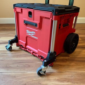 May include: Red and black rolling tool box with the "Milwaukee PACKOUT" logo. The tool box has two wheels and four smaller caster wheels for easy transport. The box is made of durable plastic.