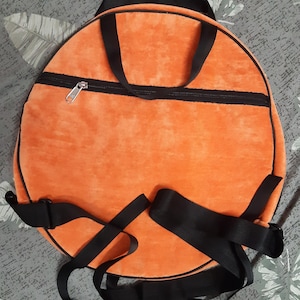May include: A round orange backpack with black straps and trim. It features a zipped pocket and a top handle. The straps are adjustable. The material looks like a soft, plush fabric.