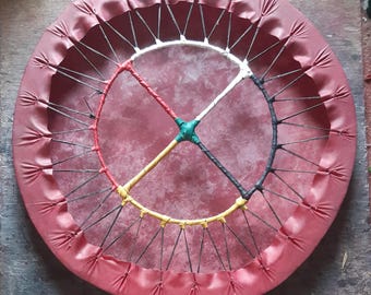 May include: A red, round drum with a white, black, green, and yellow design. The drum is made of leather and has a wooden frame.