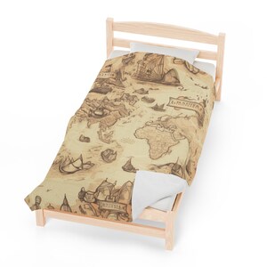 Puede incluir: Una cama individual con una almohada blanca y una funda nórdica marrón y beige con un mapa del mundo vintage con barcos y las palabras "L'IMAGINAIRE" y "SAMERICA".