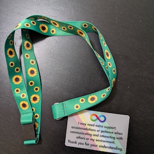 May include: A green lanyard with a sunflower pattern and a black clip. A white card with a rainbow border and an infinity symbol reads "I may need extra support, accommodations or patience when communicating and interacting with others or my environment. Thank you for your understanding."