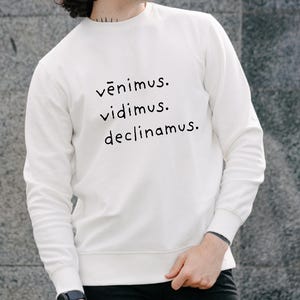 Może przedstawiać: Biała bluza z nadrukiem łacińskiego wyrażenia "Vēnimus. Vidimus. Declīnamus." w czarnym kolorze. Bluza ma długie rękawy i okrągły dekolt. Osoba na zdjęciu ma na sobie czarne spodnie i zegarek.