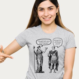 May include: A light gray t-shirt featuring a black and white graphic of two figures with speech bubbles. One bubble says, "I thought Latin was a dead language?" The other says, "No way! It's still Roman around!"