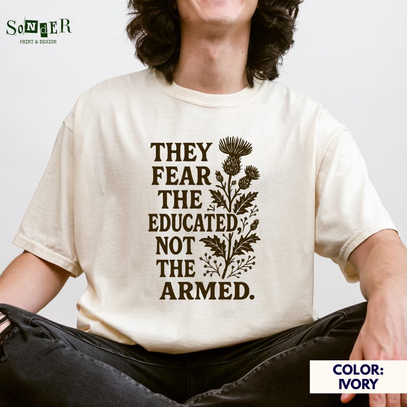 May include: A person wearing a light ivory t-shirt with a brown graphic print that reads "They fear the educated not the armed." The graphic also includes a stylized floral design.