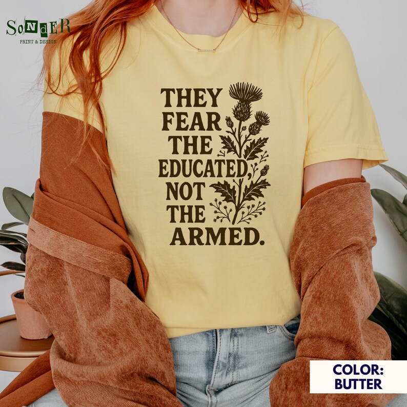 May include: A yellow t-shirt with a brown graphic print of a floral design and the text "They fear the educated, not the armed."
