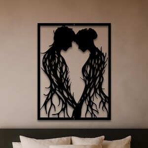 May include: Black metal wall art depicting two figures facing each other, their bodies intertwined with tree-like branches. The figures are framed within a black rectangular border, creating a silhouette effect. The artwork is mounted on a neutral-colored wall above a bed.