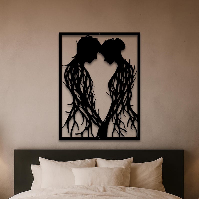 May include: Black metal wall art depicting two figures facing each other, their bodies intertwined with tree-like branches. The figures are framed within a black rectangular border, creating a silhouette effect. The artwork is mounted on a neutral-colored wall above a bed.