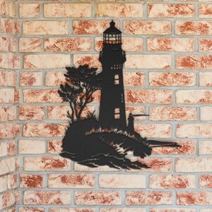 May include: A black metal wall art piece featuring a silhouette of a lighthouse standing on a rocky outcrop with a tree. The design includes detailed cutouts for the lighthouse windows and lantern room, creating a stark contrast against the wall.