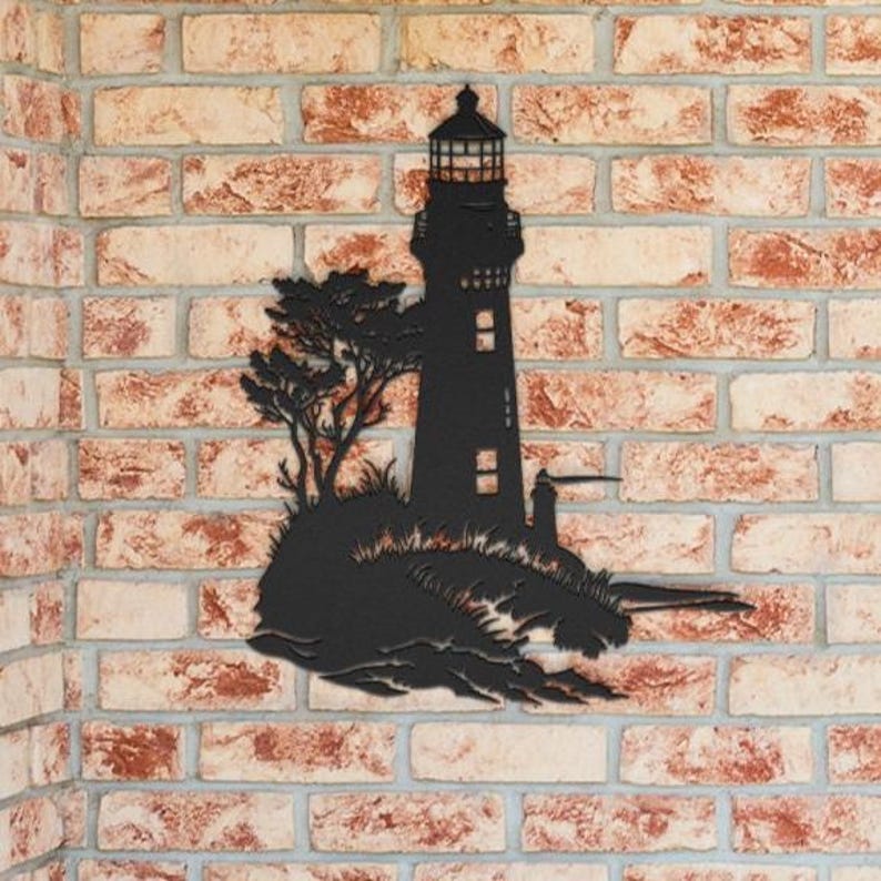 May include: A black metal wall art piece featuring a silhouette of a lighthouse standing on a rocky outcrop with a tree. The design includes detailed cutouts for the lighthouse windows and lantern room, creating a stark contrast against the wall.