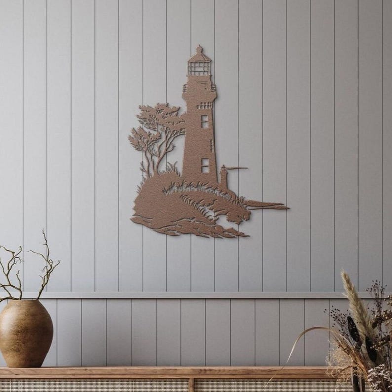 May include: A flat, copper-coloured metal wall art piece depicting a lighthouse standing on a rocky cliffside with trees. The design features a tall lighthouse structure with a lantern room at the top, set against a textured, coastal landscape.
