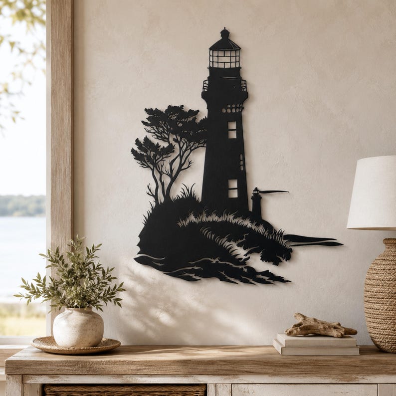 May include: A black metal wall art piece featuring a silhouette of a lighthouse standing on a rocky cliffside with a tree and crashing waves. The design includes a smaller secondary lighthouse structure and detailed cutouts for windows and the lantern room.
