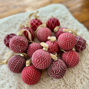 Pu&ograve; includere: Una collezione di palline di Natale rosse con intricati disegni bianchi. Le palline sono disposte su una superficie bianca e soffice.