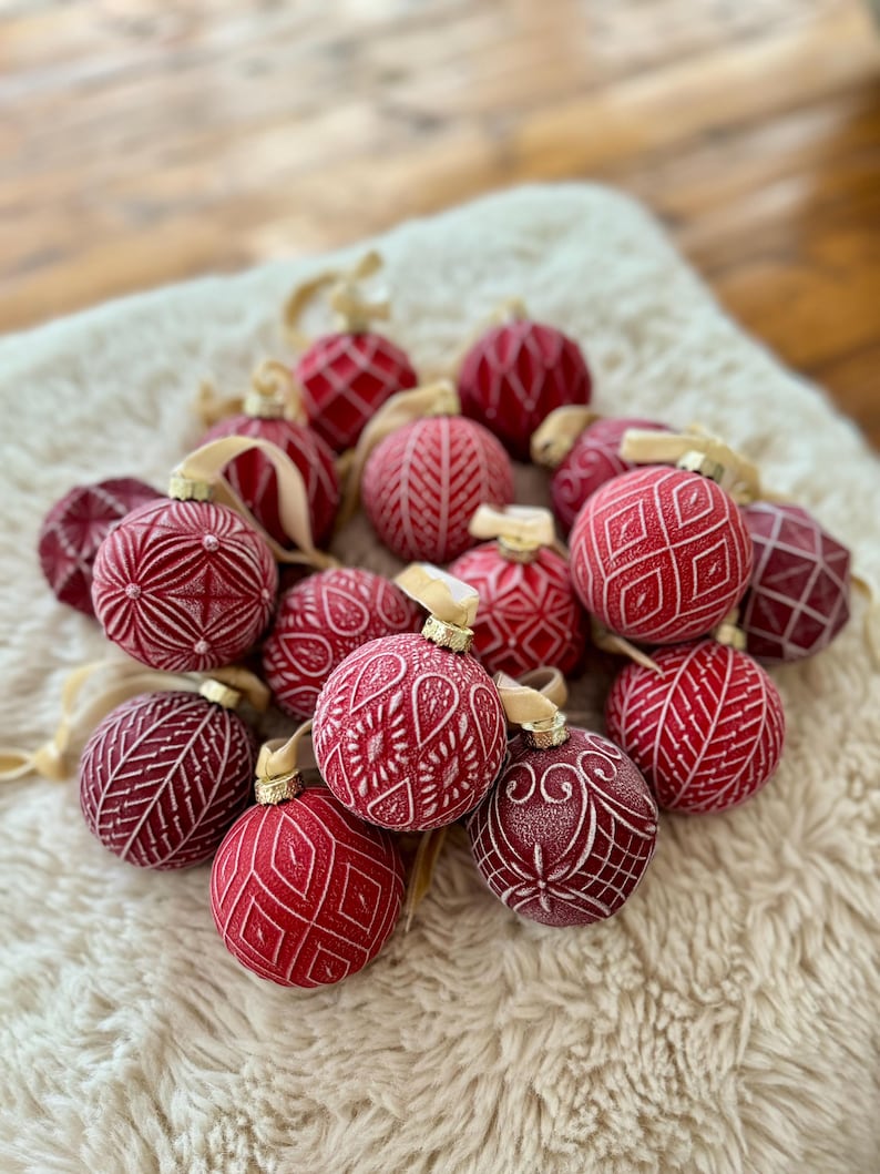 Pu&ograve; includere: Una collezione di palline di Natale rosse con intricati disegni bianchi. Le palline sono disposte su una superficie bianca e soffice.